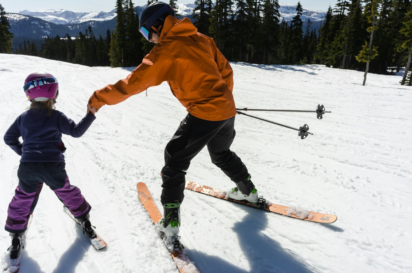A parent holding a young child's hand as they ski together on a sunny slope